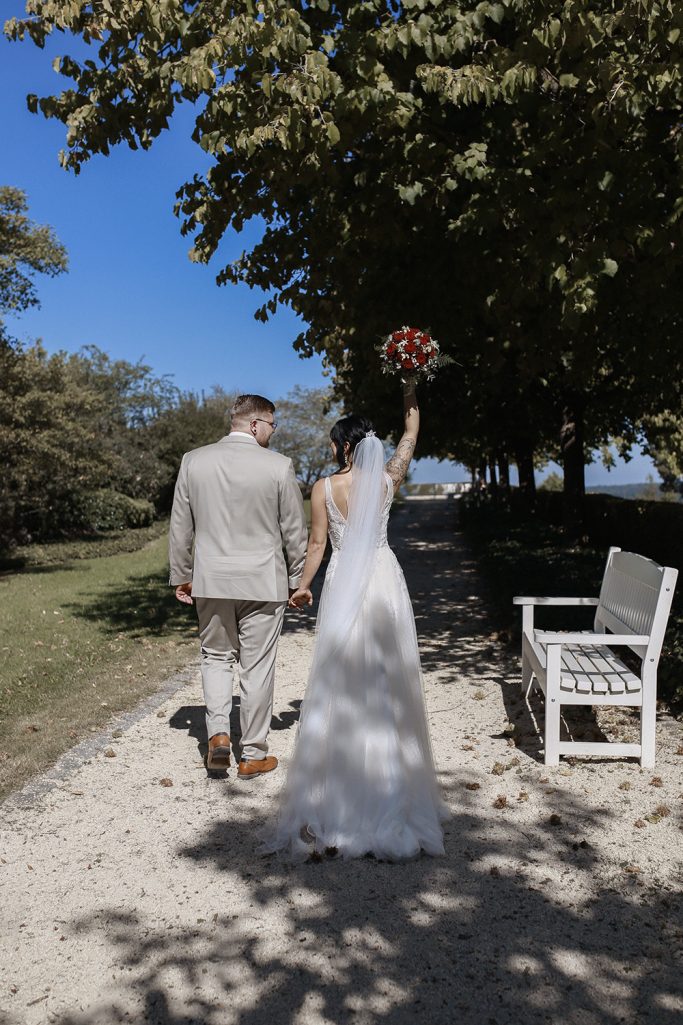 Ein Brautpaar hält Händchen, schaut sich gegenseitig liebevoll an und geht einen Weg mit Bäumen. Die Braut hält einen Blumenstrauß hoch.