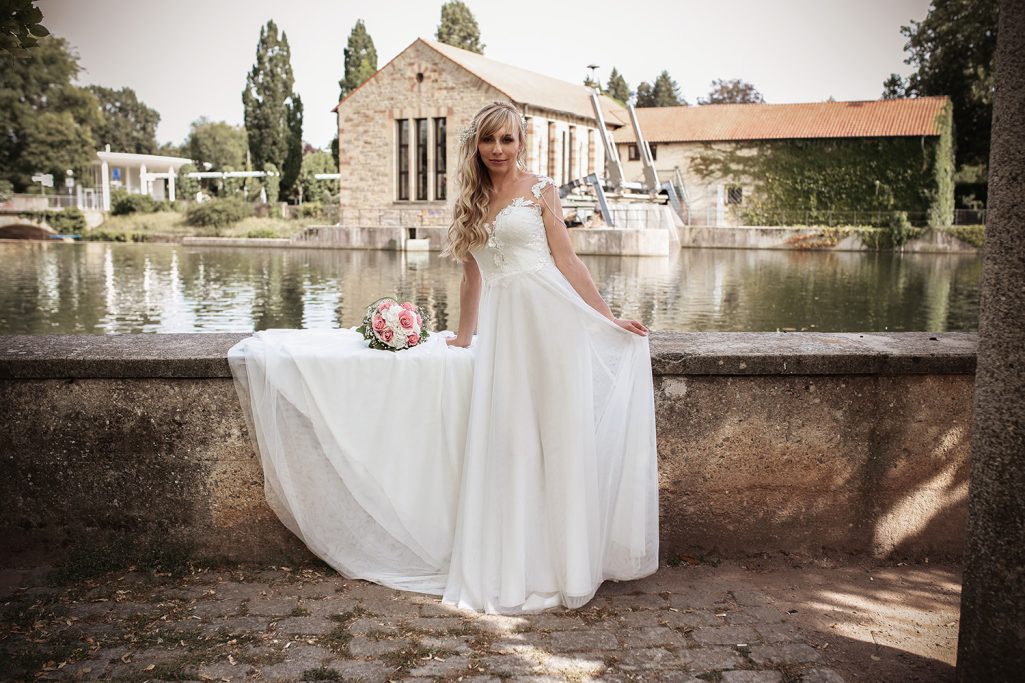 Braut in elegantem Kleid posiert am Ufer eines ruhigen Gewässers.