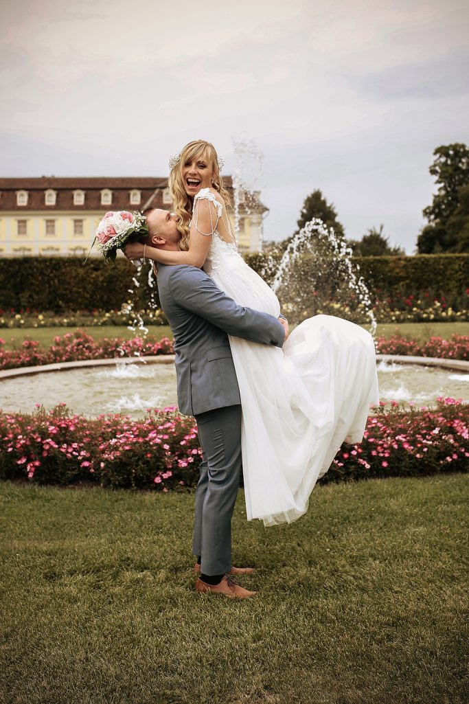 Braut und Bräutigam posieren vor einem Blumengarten mit Brunnen. Bräutigam hebt die Braut hoch, sie lächelt dabei.