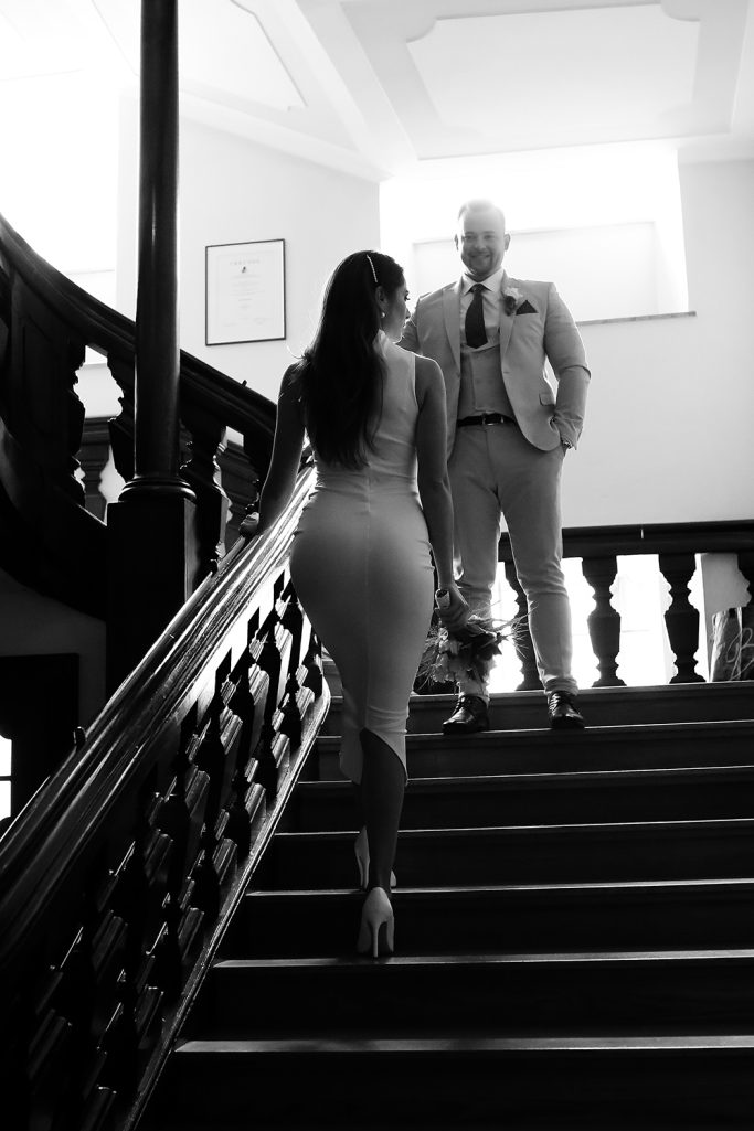 Die Braut in einem figurbetonten Kleid geht eine Treppe hinauf. Der Bräutigam wartet lächelnd auf sie und schaut sie dabei an.