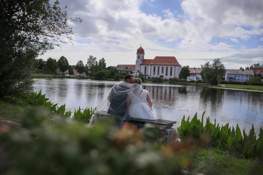 Brautpaar sitzt auf einer Bank am Ufer eines Sees mit einem historischen Gebäude im Hintergrund.