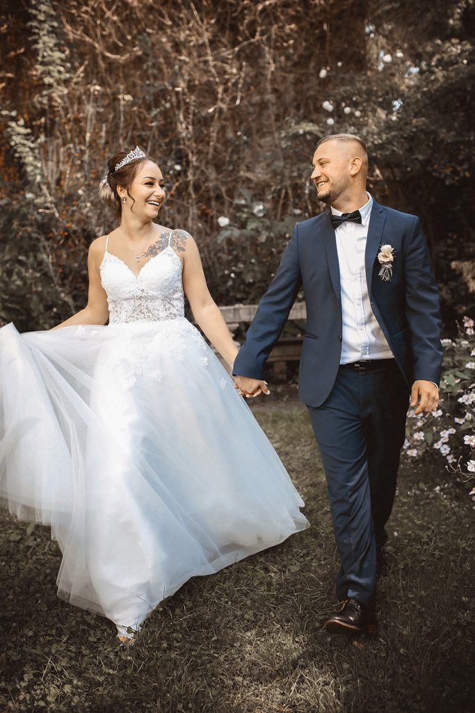 Brautpaar in eleganter Hochzeitskleidung, Hand in Hand bei Sonnenlicht in einer Gartenumgebung.