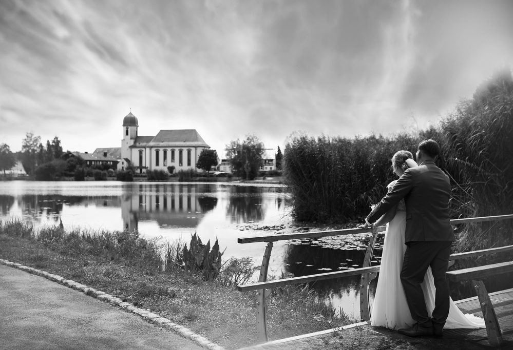 Brautpaar steht an einem See, im Hintergrund eine Kirche und bewaldete Ufer.