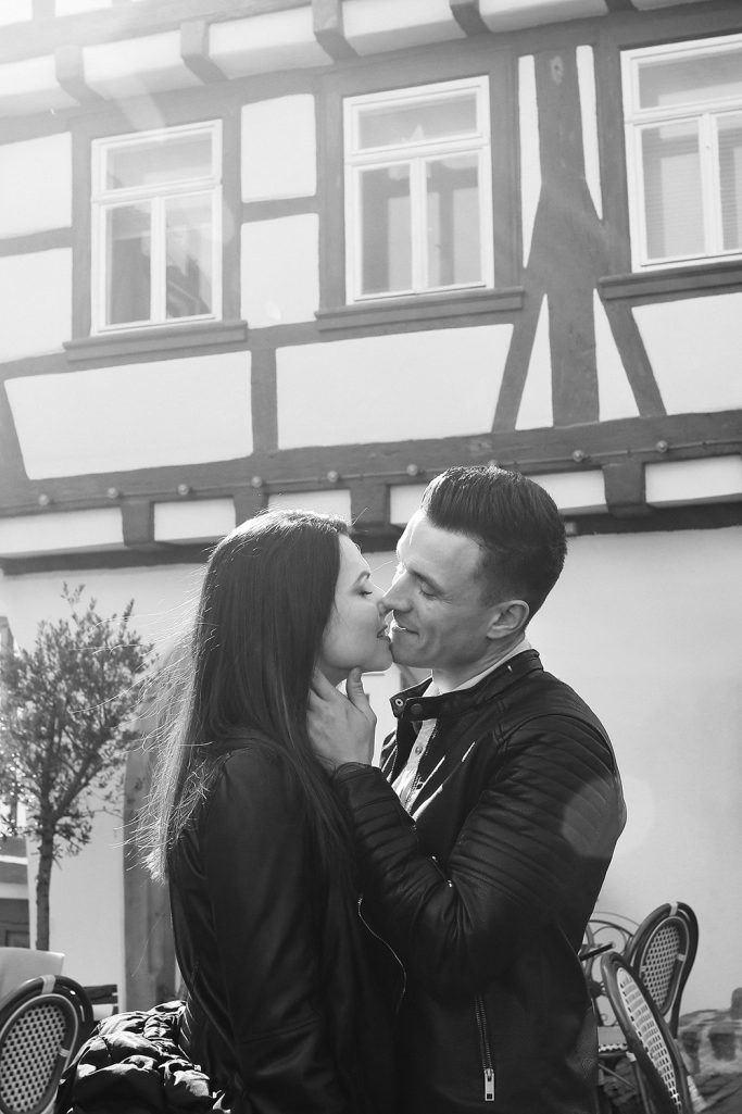 Ein Paar lächelt sich an und küsst sich vor einem Gebäude im Hintergrund.