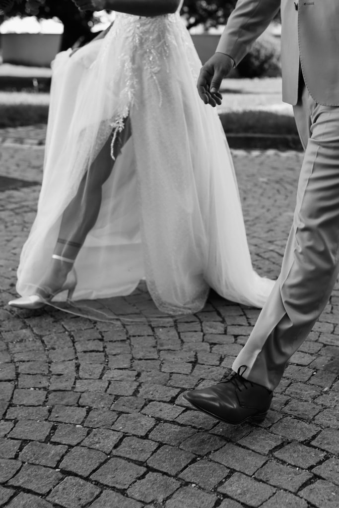 Braut in einem weißen Kleid mit Schleppe, neben ihrem Bräutigam. Beide laufen gemeinsam einen gepflasterten Weg entlang.