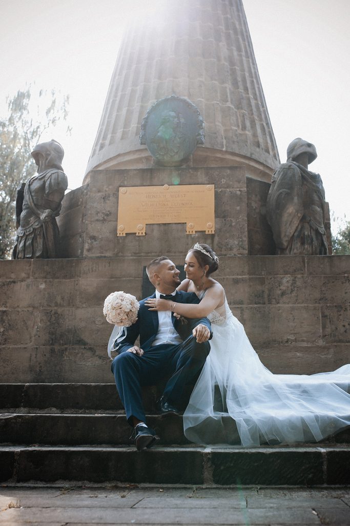 Brautpaar mit Blumenstrauß sitzt auf Stufen vor einem monumentalen Denkmal im Freien.