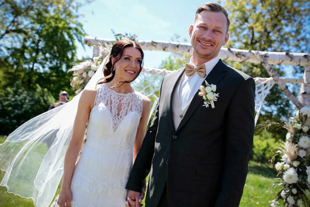 Braut und Bräutigam lächeln glücklich und posieren vor einer grünen Landschaft im Freien.