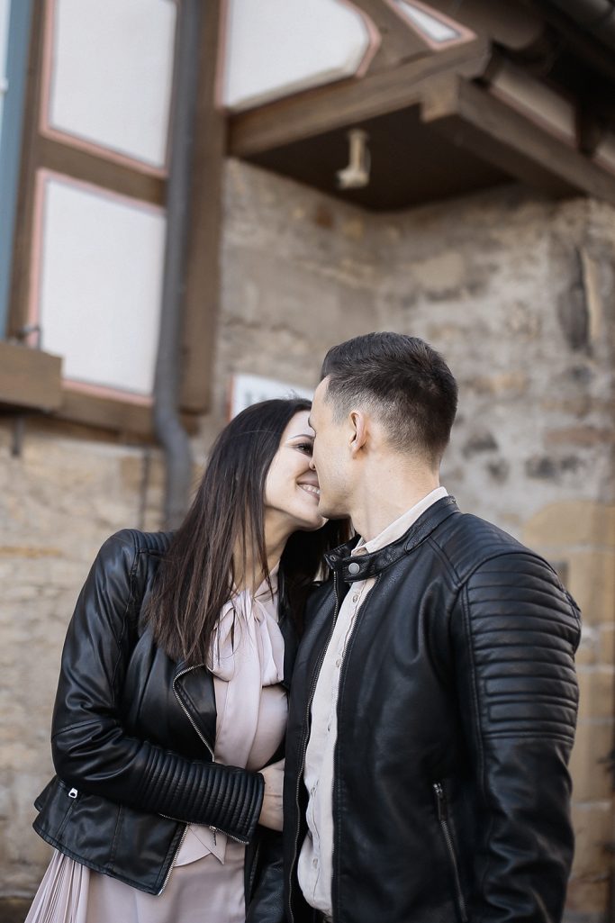 Paar in Umarmung, küsst sich vor einer Steinmauer. Sie tragen modische Jacken.