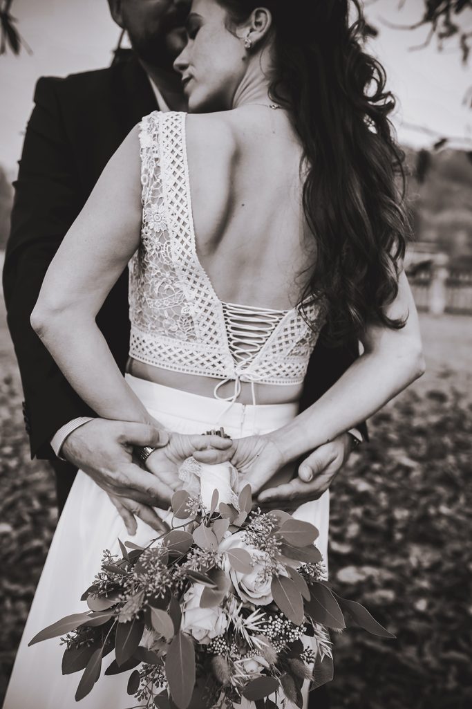 Braut in einem eleganten Kleid umarmt von ihrem Partner, mit Blumenstrauß in der Hand.