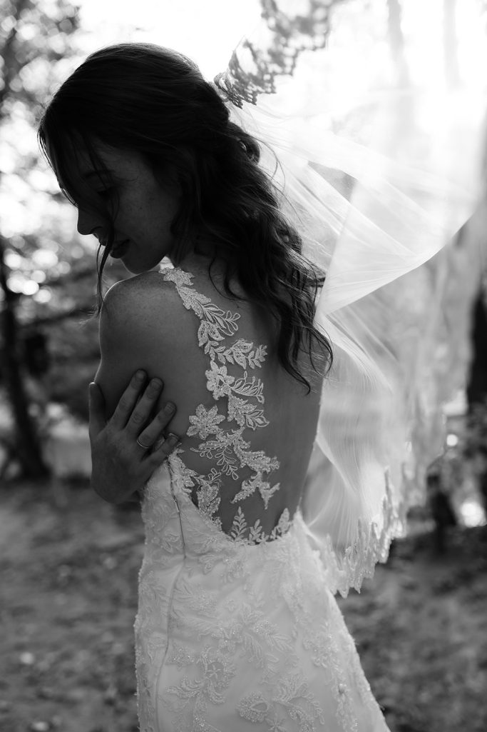 Braut mit offenem Rücken in einem fließenden weißen Kleid und langem Schleier. Schaut emotional nach links über die eigene Schulter.