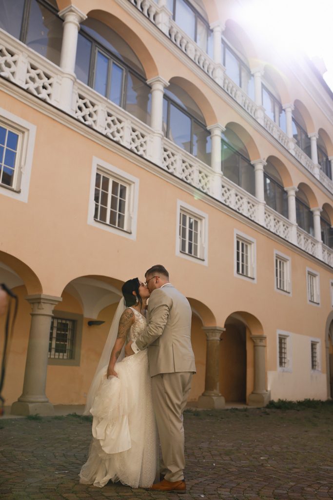 Ein Brautpaar küsst sich romantisch vor einem historischen Gebäude mit Bogenfenstern.