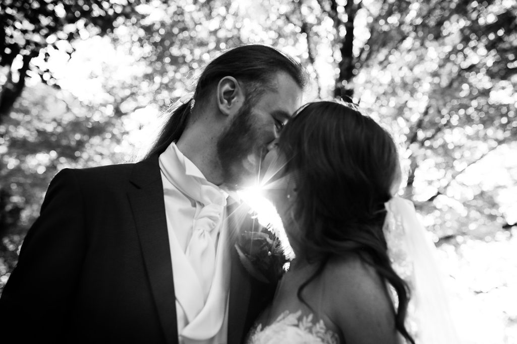Brautpaar küsst sich im Freien, umgeben von Bäumen, mit Sonnenlicht im Hintergrund.