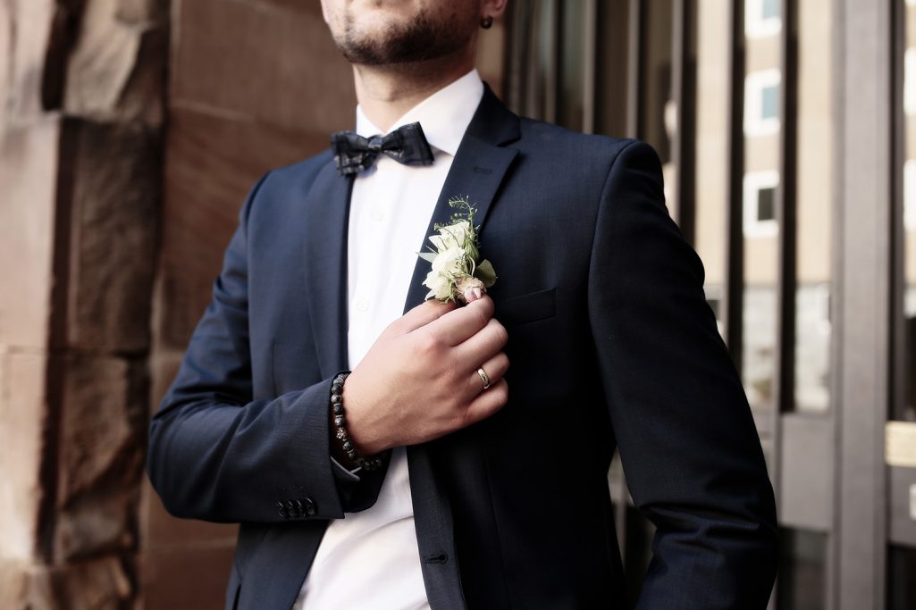 Bräutigam in festlichem Anzug mit Fliege und Anstecker, elegant posierend.