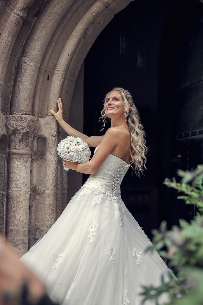 Braut mit langem, blondem Haar in einem eleganten weißen Kleid, hält einen Blumenstrauß.