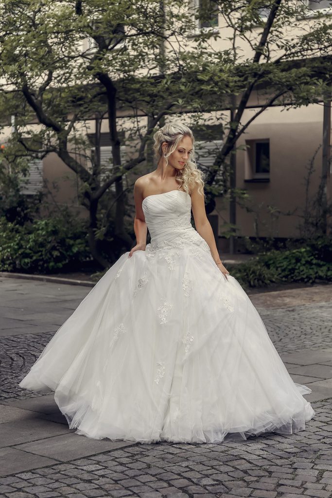 Braut in Brautkleid posiert auf gepflastertem Weg. Hinter ihr ein Gebäude mit Bäumen davor.