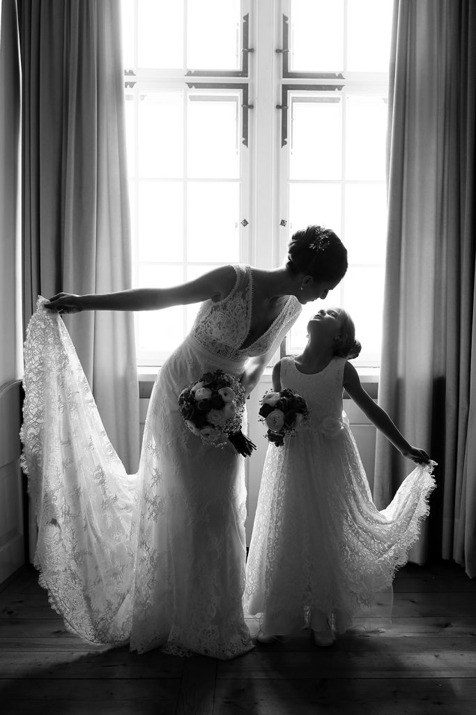 Braut und Mädchen in weißen Kleidern, die sich liebevoll anschauen und die Kleider sowie einen Blumenstrauß halten.