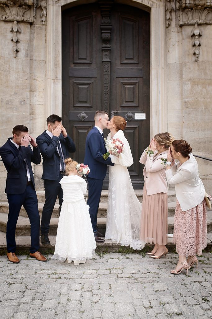 Braut und Bräutigam küssen sich, umgeben von Hochzeitsgästen auf einer Treppe, die ihre Augen mit den Händen verschließen.