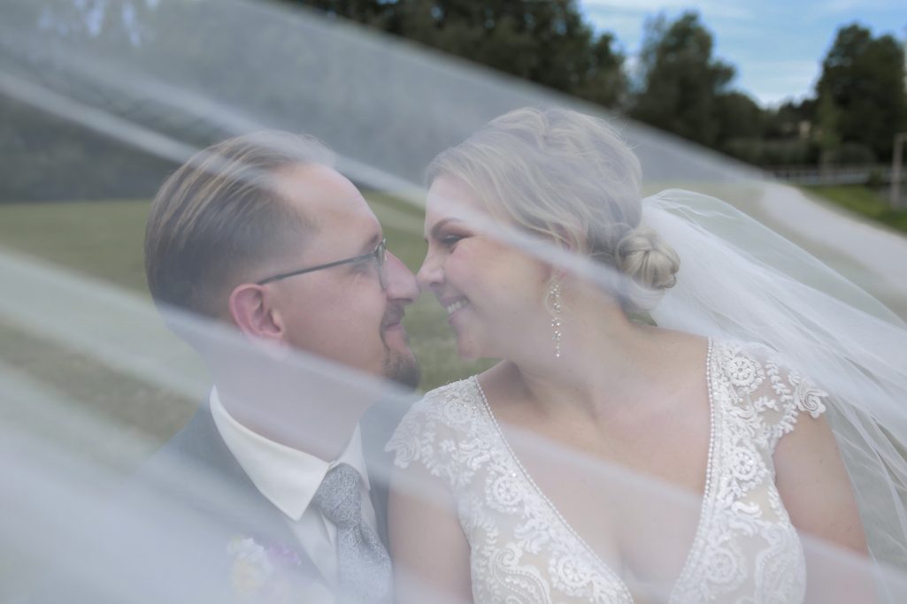 Brautpaar lächelt sich gegenseitig liebenswürdig an, unter einem transparenten Schleier in der Natur.