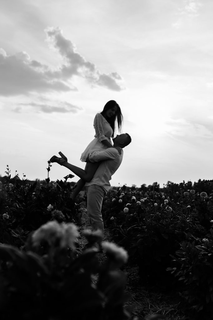 Paar steht in einem Feld. Mann hebt Frau in weißem Kleid vor einem Sonnenuntergang.