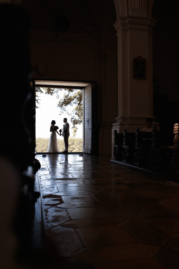 Brautpaar steht in einem abgedunkelten Kirchenraum vor einem hellen Gebäudeausgang.