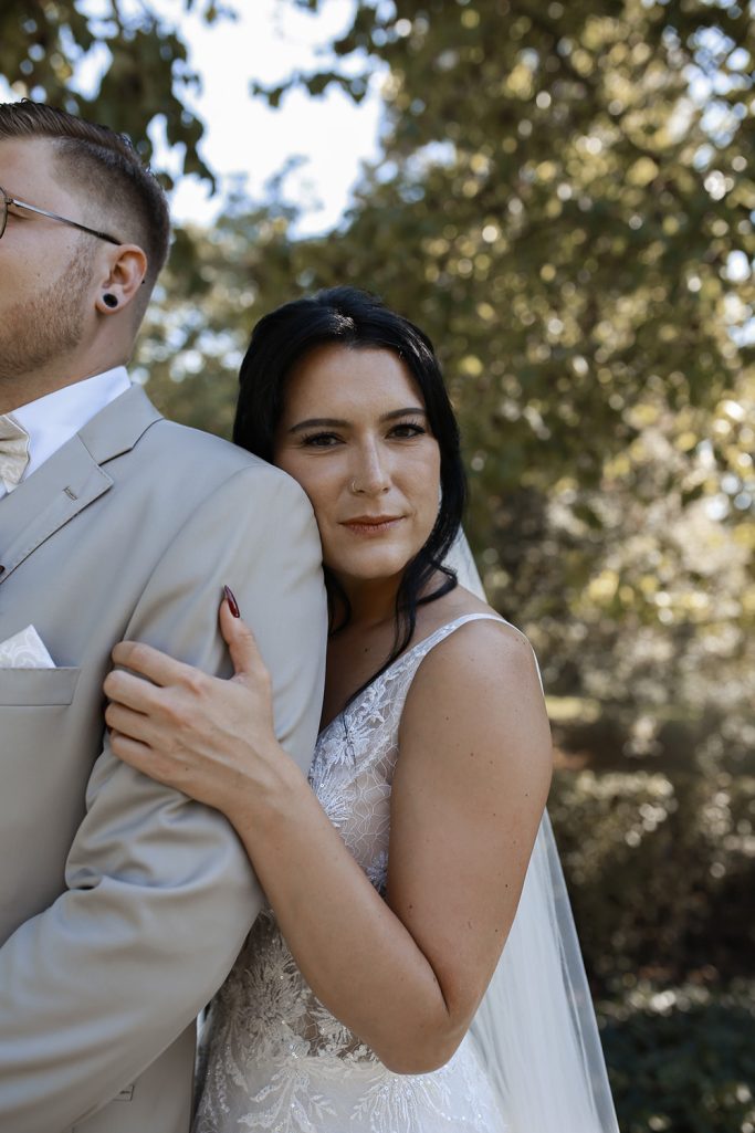 Braut umarmt den Bräutigam, beide im Freien mit grünem Hintergrund.