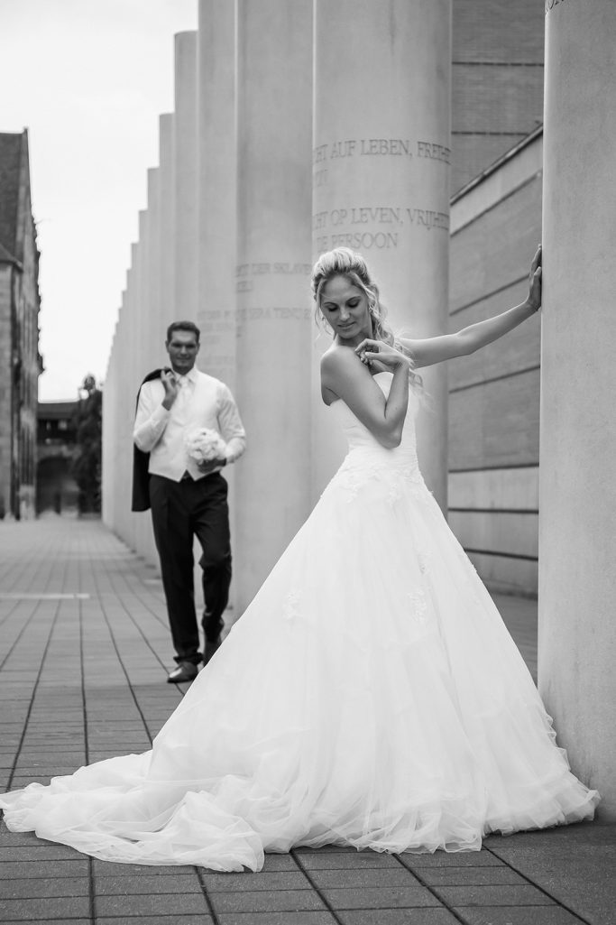 Braut in einem weißen Kleid, steht an einer Säule, während der Bräutigam im Hintergrund auf sie zuläuft.