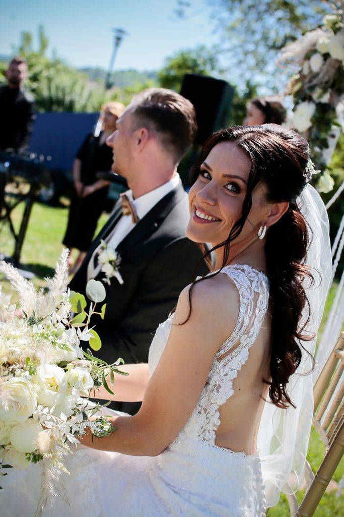 Braut in elegantem Kleid sitzt gemeinsam mit Bräutigam an einem gedeckten Hochzeitstisch.