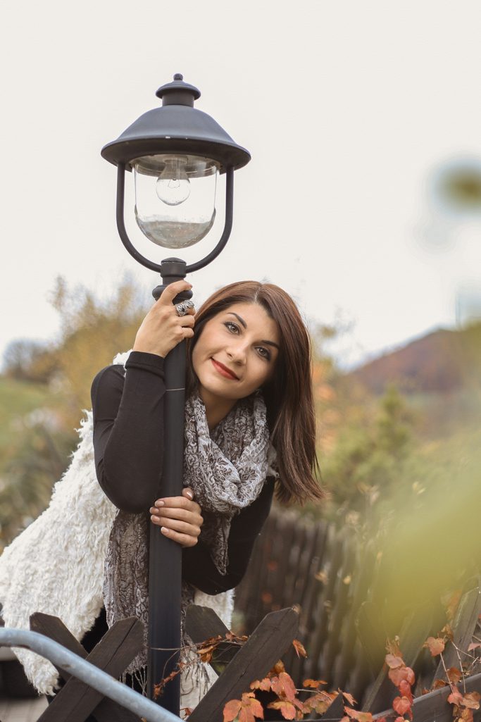 Eine Frau lehnt lächelnd an einem Laternenpfahl in einer herbstlichen Umgebung.