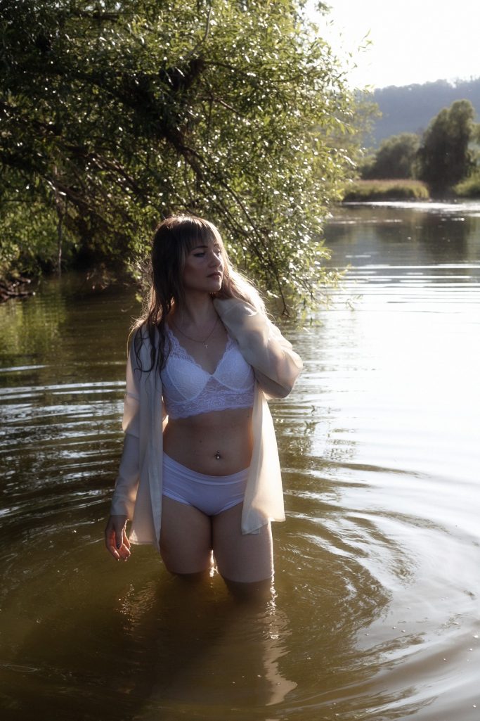 Frau im weißen Bikini steht im flachen Wasser, umgeben von Bäumen und Sonnenschein.