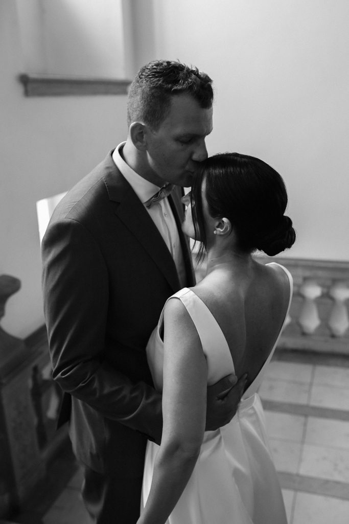 Brautpaar umarmt sich zärtlich in einem eleganten Raum, Bräutigam küsst liebevoll Stirn der Braut, Schwarz-Weiß-Fotografie.