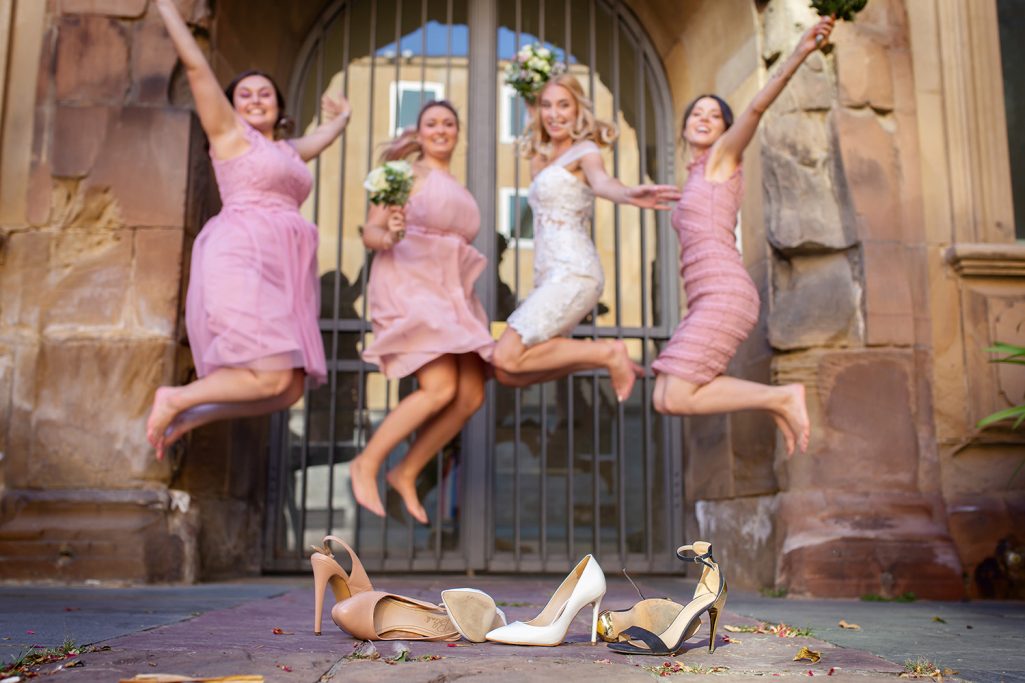Braut und drei weitere Frauen springen mit ausgezogenen Schuhen glücklich in der Luft vor einem Mauertor.