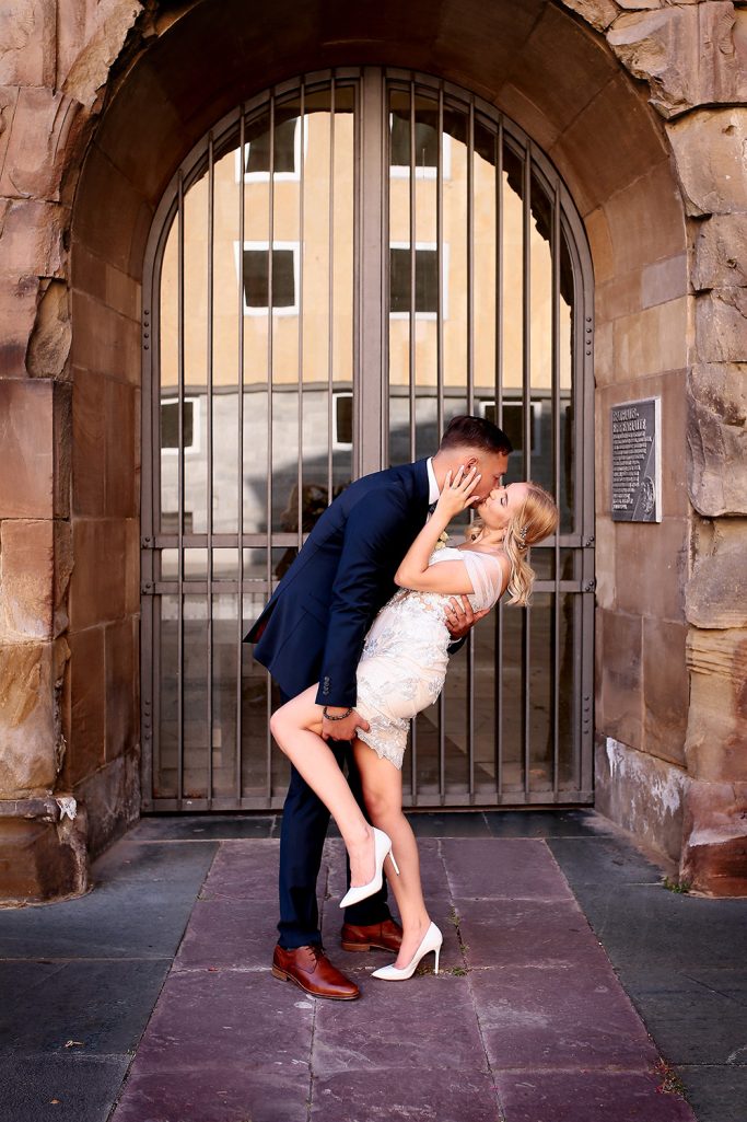 Brautpaar umarmt und küsst sich innig vor einem Tor. Beide glücklich in eleganter Kleidung und in romantischer Stimmung.