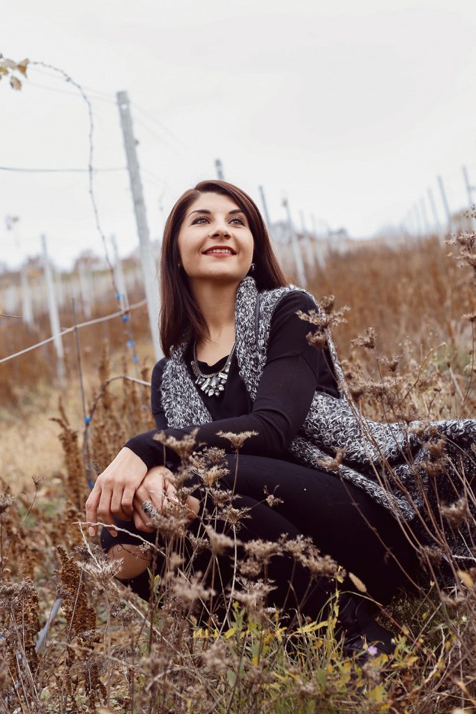 Frau mit dunklem Haar posiert in einer Wiese, lächelt und trägt einen grauen Strickmantel.