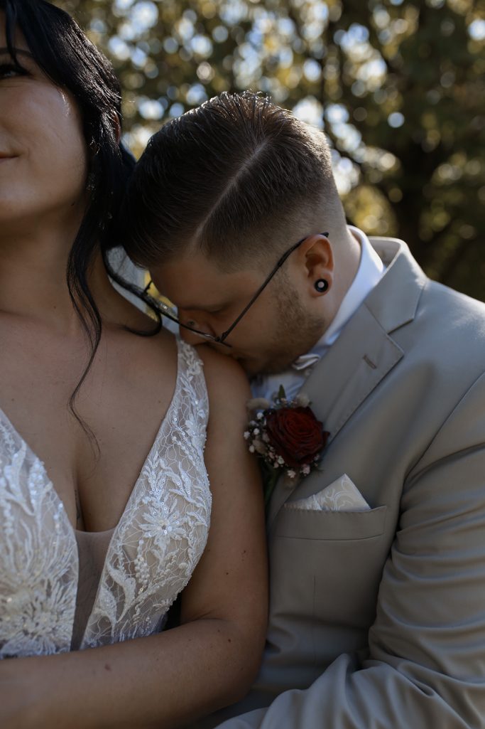 Brautpaar in eleganter Hochzeitskleidung, eng umschlungen und verliebt. Bräutigam küsst liebevoll die Schulter der Braut.