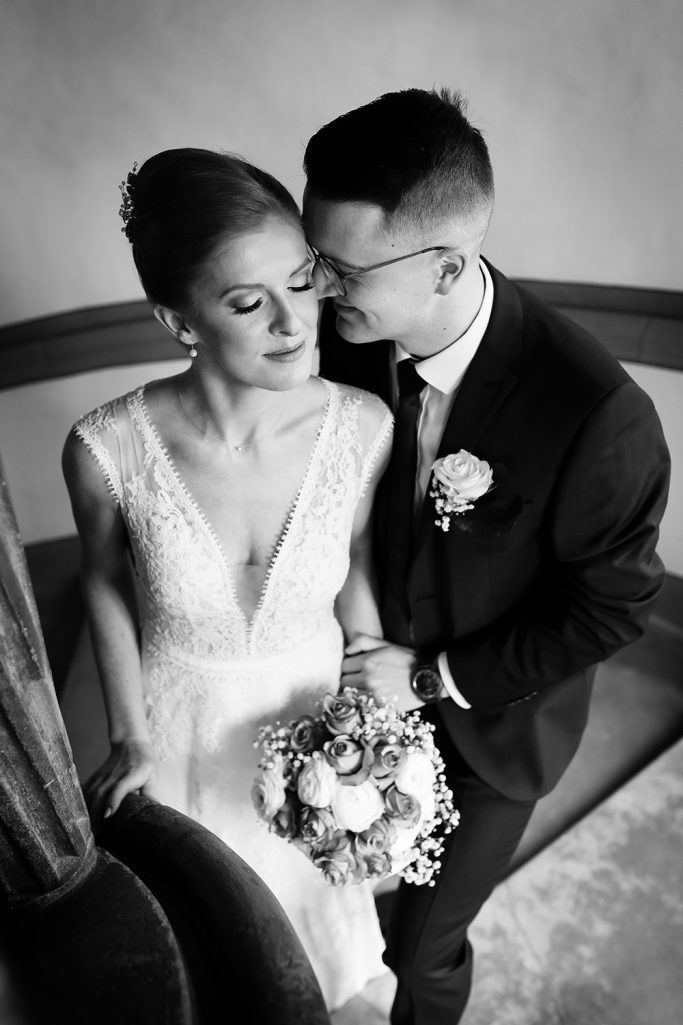 Braut und Bräutigam umarmen sich liebevoll stehend auf einer Treppe, mit einem Blumenstrauß in der Hand.