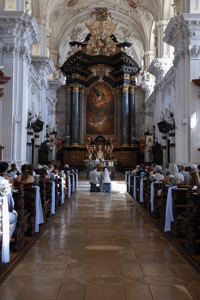 Blick auf den Altar einer Kirche mit Sitzbänken und Menschen während einer Hochzeitszeremonie.