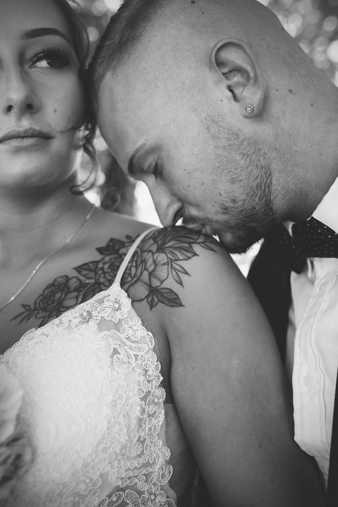 Braut in weißem Kleid und Tattoo. Bräutigam küsst liebevoll die Schulter der Braut.