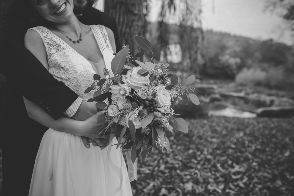 Eine Braut in einem eleganten Kleid im Freien hält einen Blumenstrauß, umarmt von ihrem Bräutigam.
