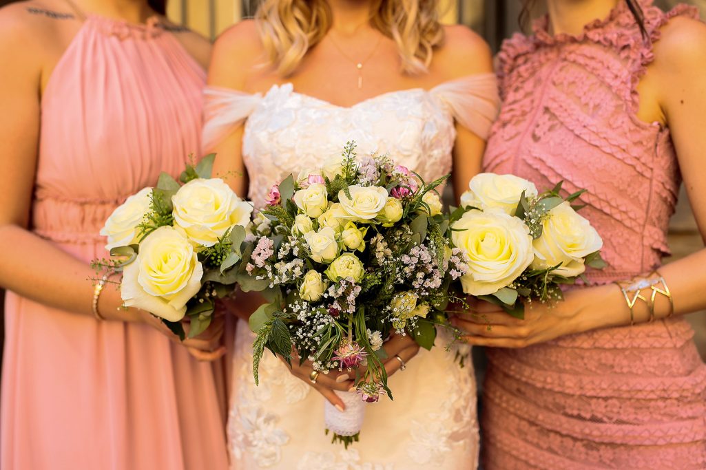 Hochzeitsblumenstrauß mit weißen Rosen und grünen Pflanzen, gehalten von der Braut und zwei weiteren Frauen.