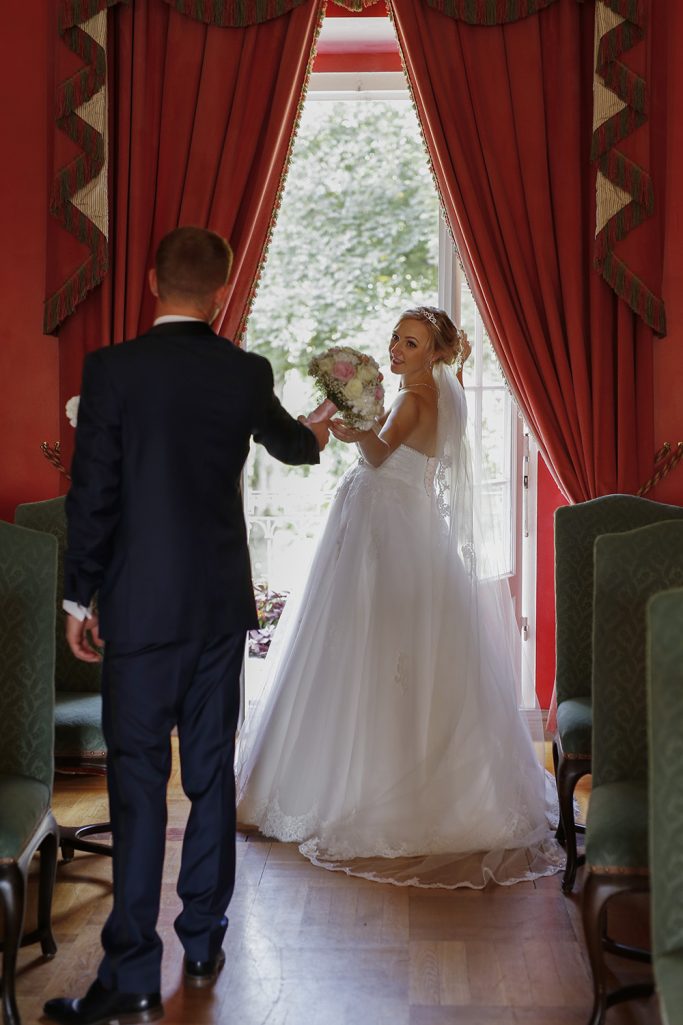 Braut in weißem Kleid steht vor einem Fenster, Bräutigam reicht ihr einen Blumenstrauß.