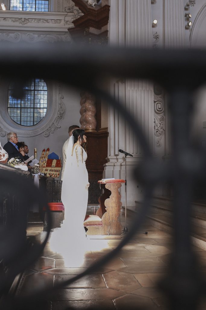Braut in weißem Kleid steht am Altar, flexible Perspektive durch Geländer sichtbar.