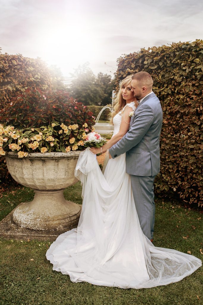 Braut und Bräutigam umarmen sich vor einem Blumenarrangement im Freien.