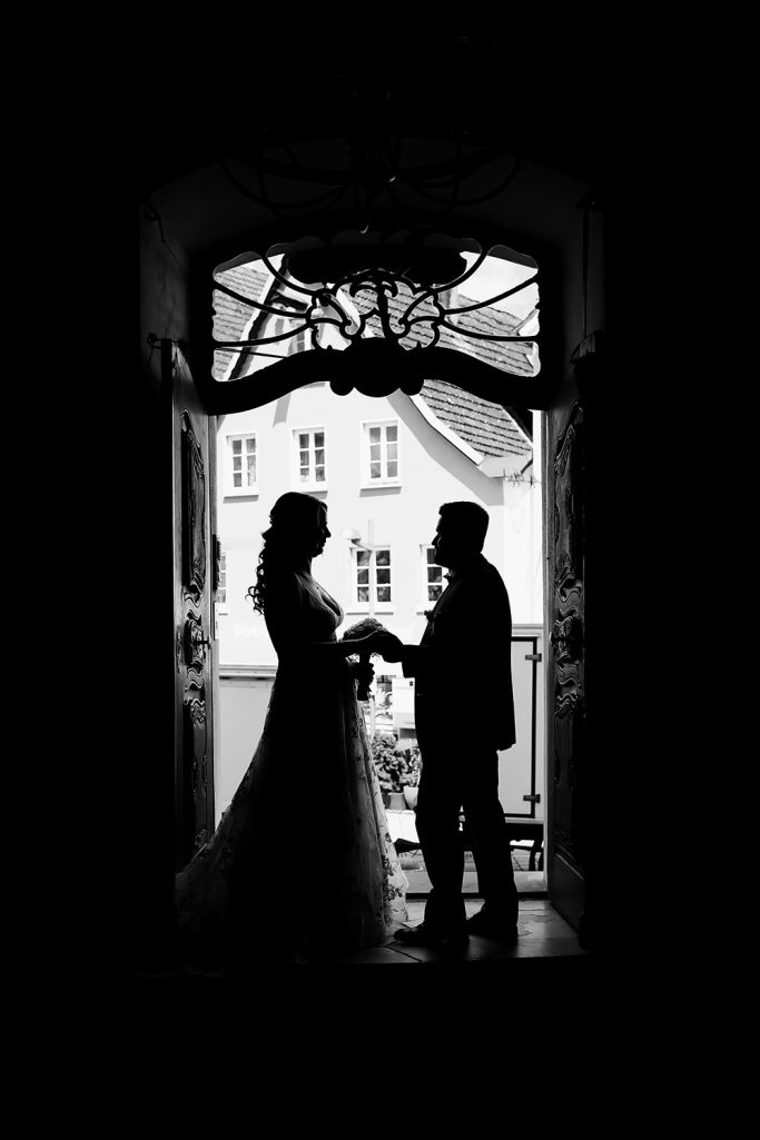 Hochzeitspaar in Silhouette, stehend im Türrahmen. Hält Händchen und schaut sich gegenseitig liebevoll an.
