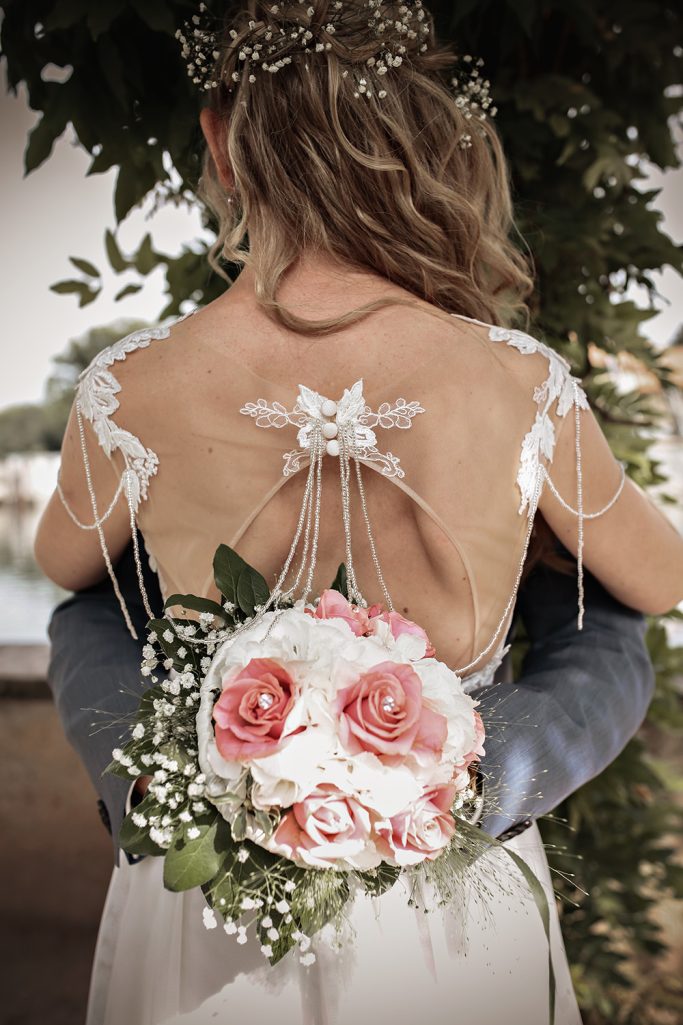 Braut mit schulterfreiem Kleid, umarmt vom Bräutigam, der einen Blumenstrauß in den Händen hält - von hinten fotografiert.