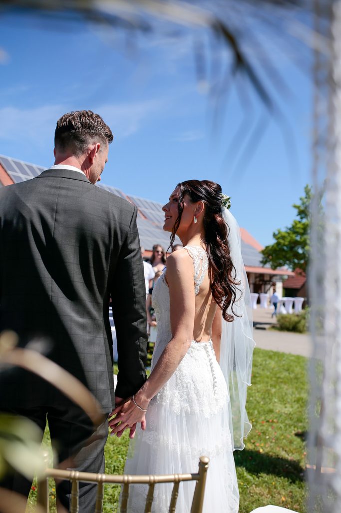 Braut und Bräutigam halten Händchen, stehen im Freien mit blauem Himmel im Hintergrund und schauen sich gegenseitig an.