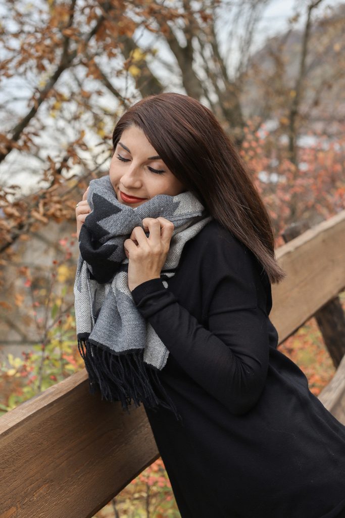 Frau mit dunklen Haaren und Schal, lächelt vor herbstlicher Kulisse.