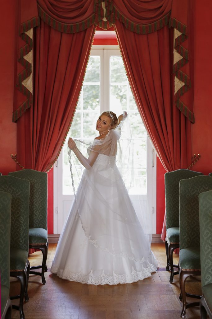 Braut in einem weißen Kleid steht vor einem großen Fenster mit roten Vorhängen.