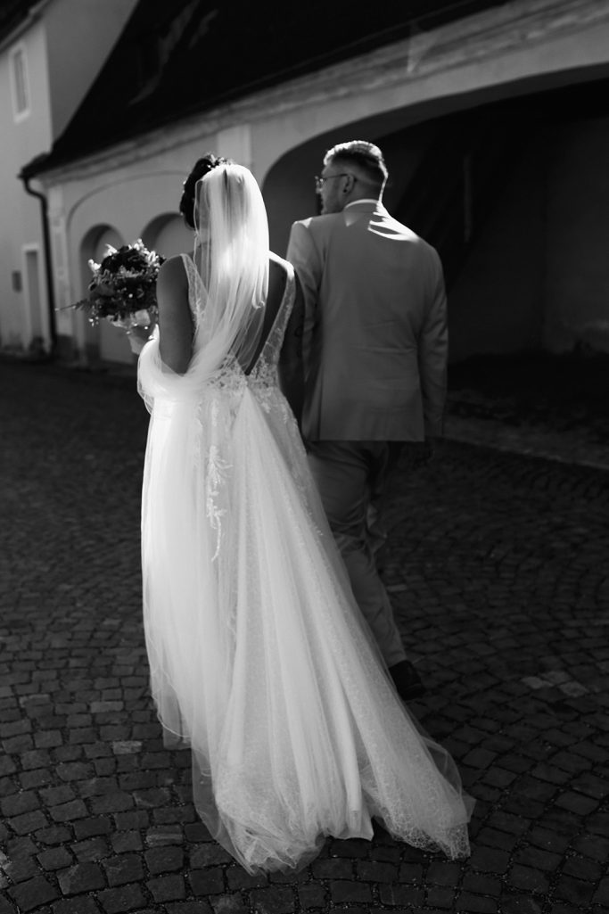 Brautpaar geht Hand in Hand einen gepflasterten Weg entlang. Der Bräutigam trägt einen hellen Anzug und die Braut ein langes weißes Kleid.