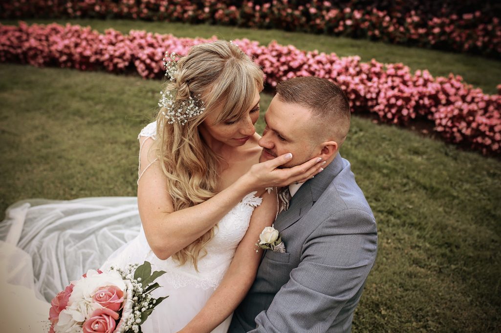 Braut und Bräutigam sitzen zusammen in einem Blumenbeet und lächeln sich an.