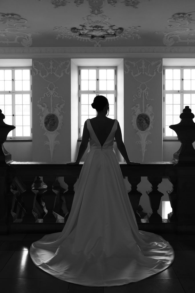 Braut in einem eleganten Kleid steht silhouettenhaft an einem Balkon mit Fenstern.